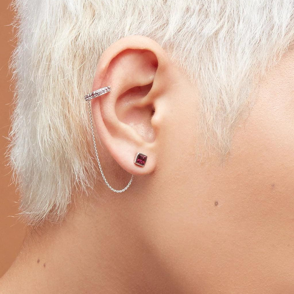 Ear with a short platinum-blonde hairstyle wearing a small ruby-red square stud earring paired with a silver ear cuff connected by a delicate chain, creating a layered, edgy look.