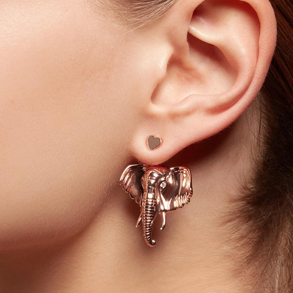Close-up of an ear adorned with a small rose gold heart-shaped stud paired with a large detailed elephant head charm in matching tone.