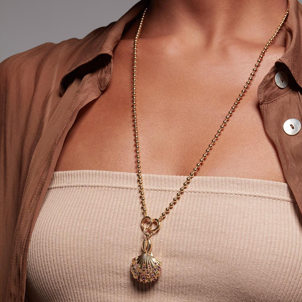 Gold ball chain necklace with shell-inspired pendant adorned in multicolor stones on a woman wearing a strapless beige top"
