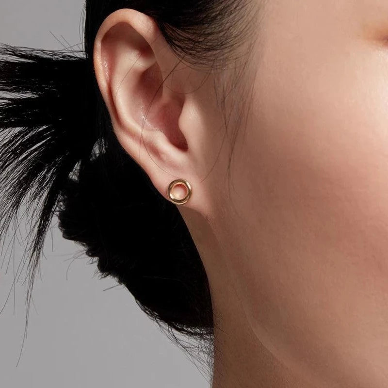 Close-up of an ear wearing a small, polished circular stud earring in gold. The earring is minimalist with an open circle design, set against a neutral background.
