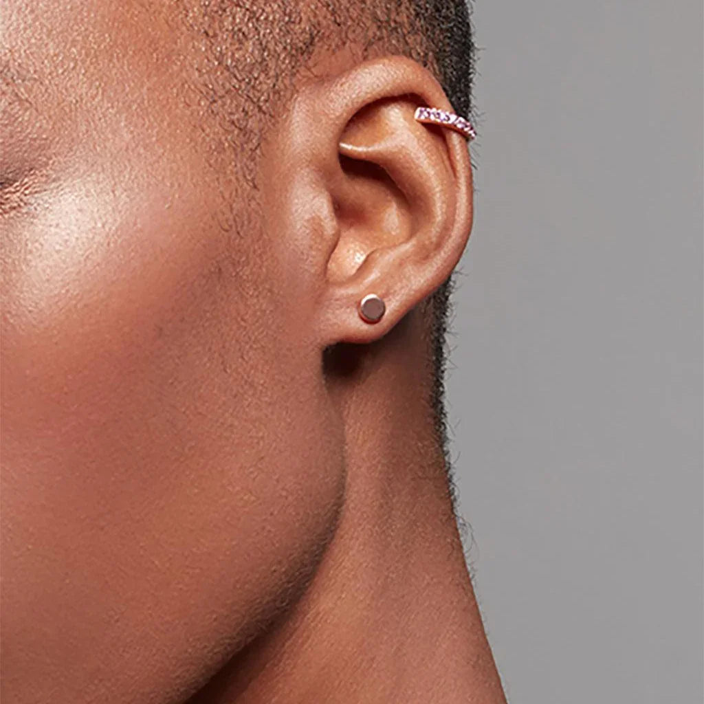 Close-up of an ear with a small, round, rose gold stud earring on the lobe and a pink gemstone ear cuff on the upper ear, worn on dark skin.