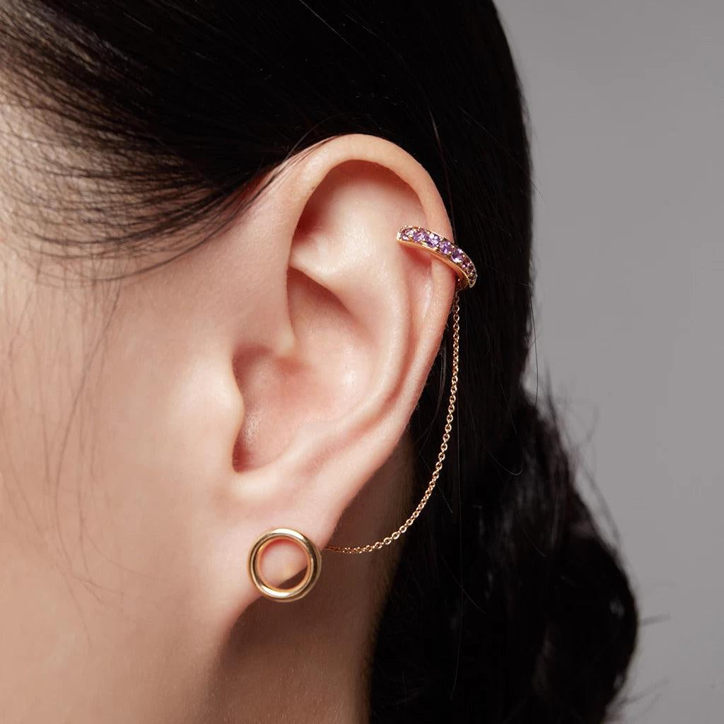 Gold ear cuff with purple stones and attached chain styled with a circular gold stud on a light-skinned ear.