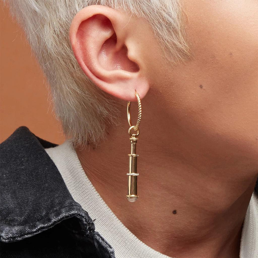 Gold-tone twisted hoop earring with telescope-shaped pendant worn on ear.