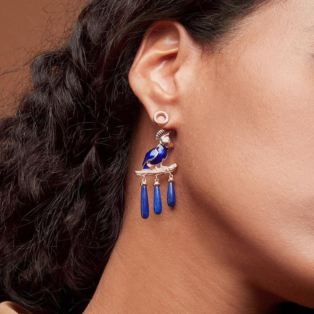 Detailed shot of an ear with a rose gold circular stud used as a base for an elaborate dangling bird charm. The charm features a blue enamel bird perched on a branch with three blue teardrop-shaped dangles.