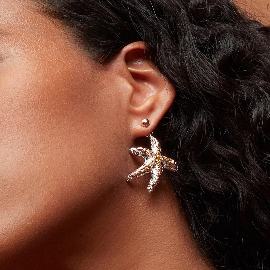 A close-up of an ear wearing a small round ball stud with a large dangling starfish-shaped charm attached. The starfish is textured and metallic with intricate detail.