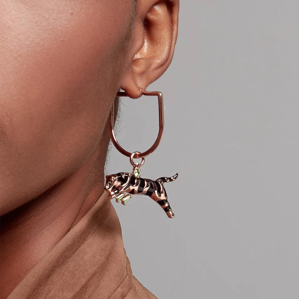 Close-up of a woman's ear wearing a rose gold geometric hoop earring with a black and gold striped leaping leopard charm featuring green gemstone eyes.