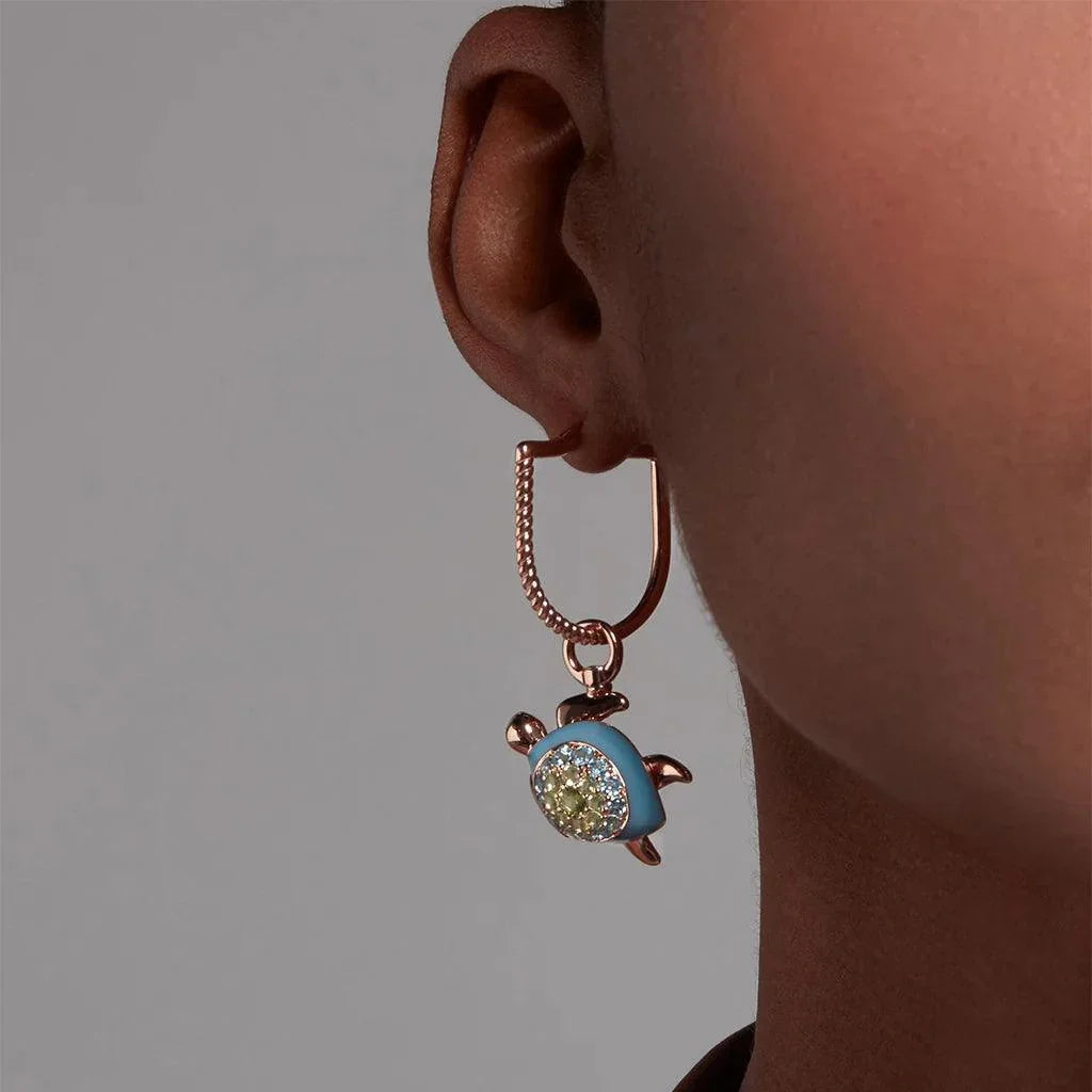 Close-up of an ear wearing a rose gold square hoop earring with a dangling turtle charm featuring a blue enamel shell and sparkling crystal center.