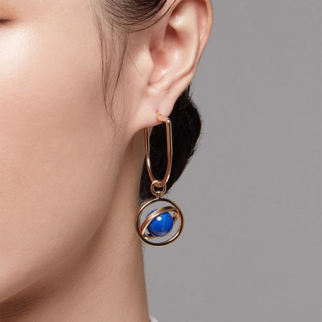 Close-up of a woman's ear wearing a gold hoop earring with a dangling charm designed like a blue planet encircled by a gold ring.