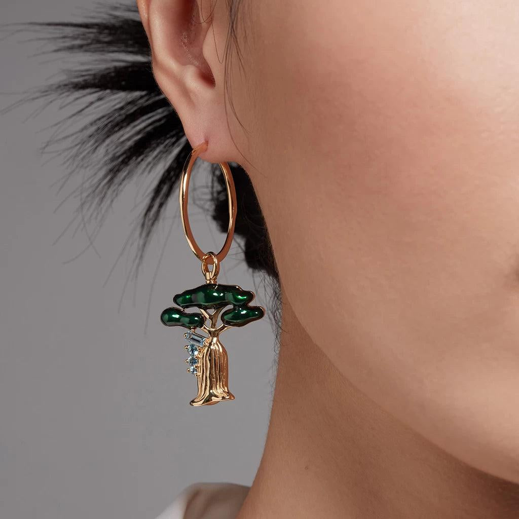 Close-up of a woman's ear wearing a gold round hoop earring with a dangling charm shaped like a baobab tree, featuring green enamel leaves and blue gemstones.