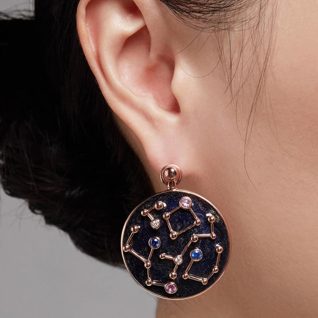 Close-up of an ear wearing a small ball stud with a large round drop charm featuring a navy blue background and rose gold constellation designs with colored stones.