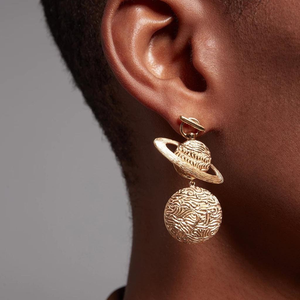 Close-up of an ear wearing gold drop earrings featuring a textured Saturn-shaped charm with rings above a detailed spherical charm, set against dark skin.
