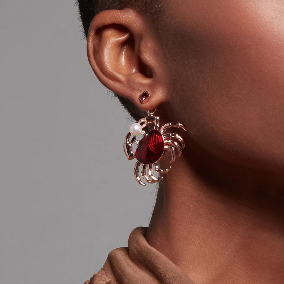 Dark-skinned model wearing a ruby-red square stud earring with a large, dramatic crab-shaped dangle charm featuring red enamel and a pearl accent.