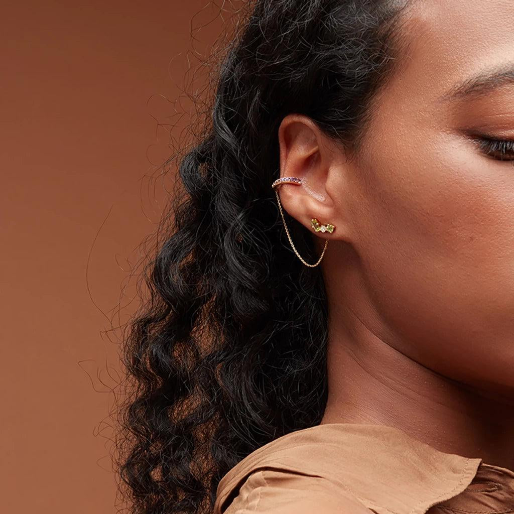 Gold chain-link ear cuff with purple stones worn on curly-haired model, styled with green gemstone stud below.