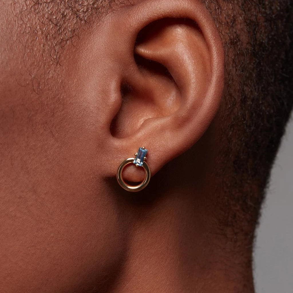 Close-up of ear with small gold hoop stud featuring rectangular blue gemstone.