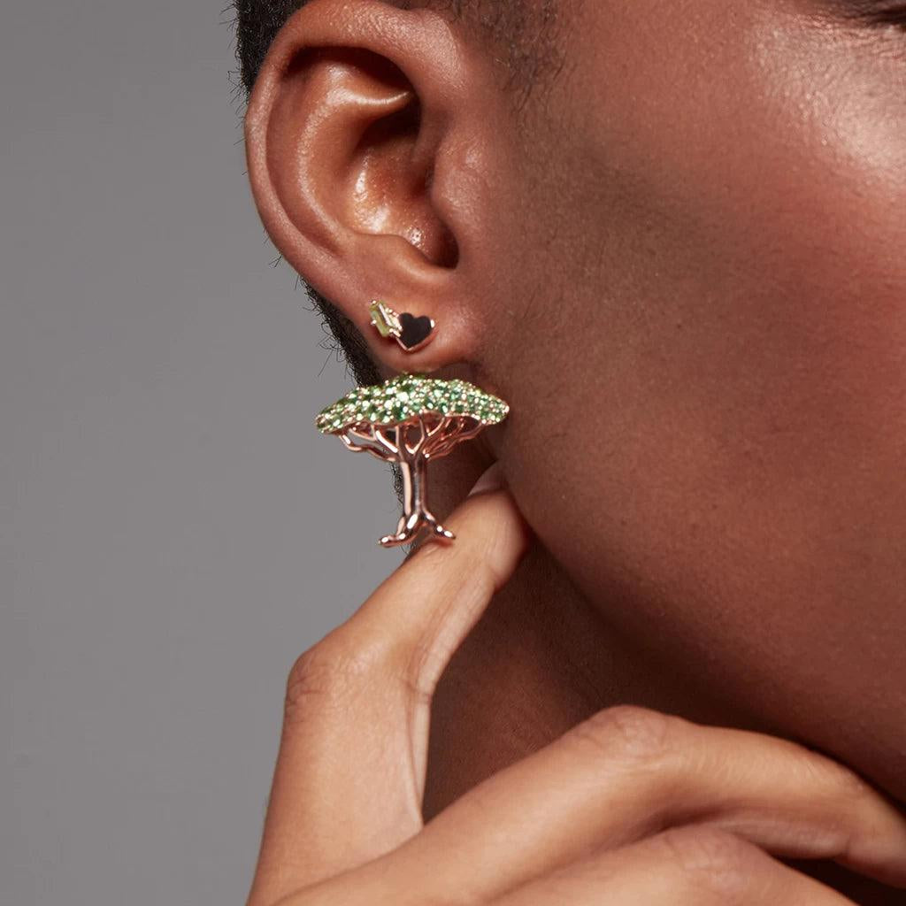 Statement heart-shaped earring with green stones paired with tree-shaped charm worn on ear.