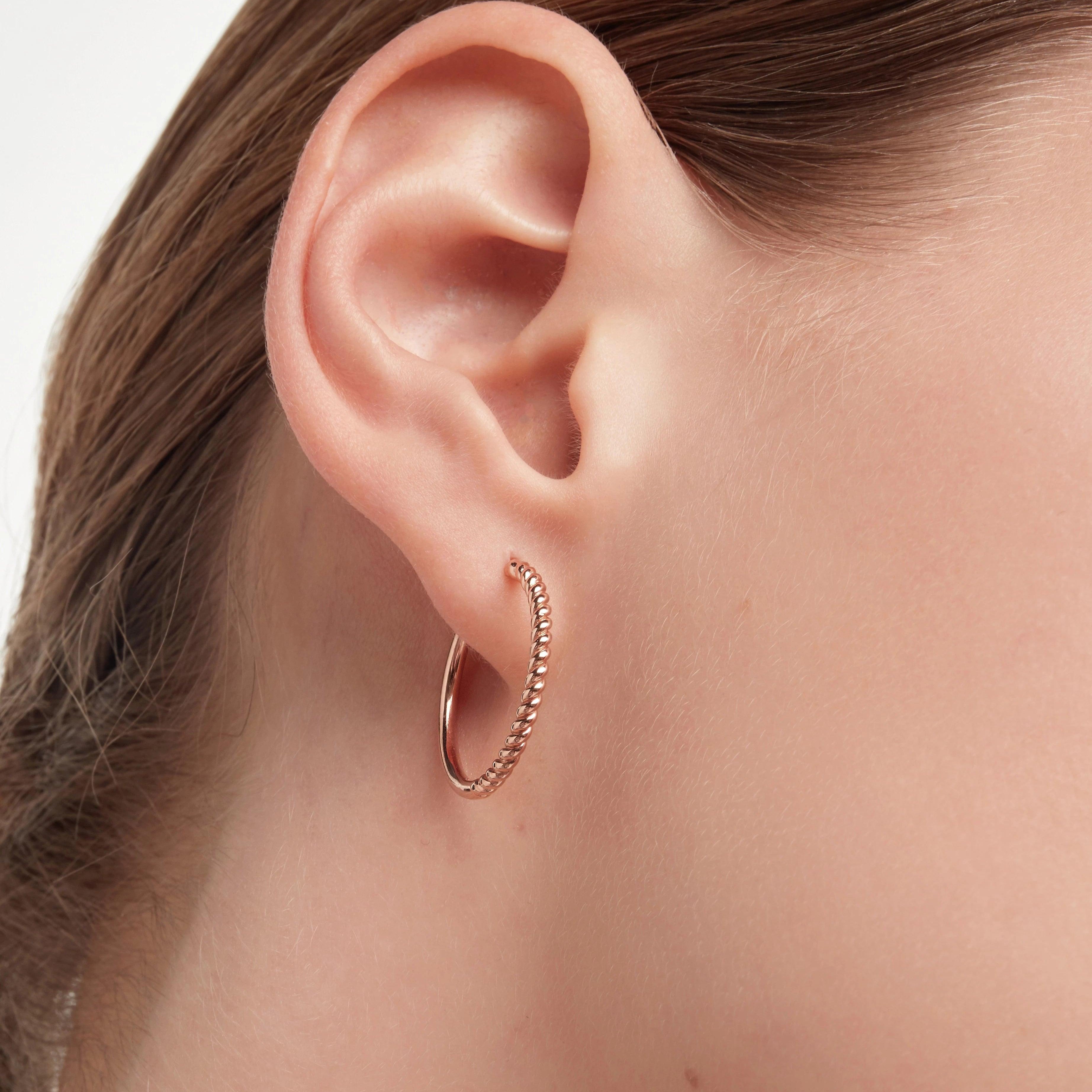 Plain rose gold-tone twisted hoop earring with twisted rope detailing worn on ear.