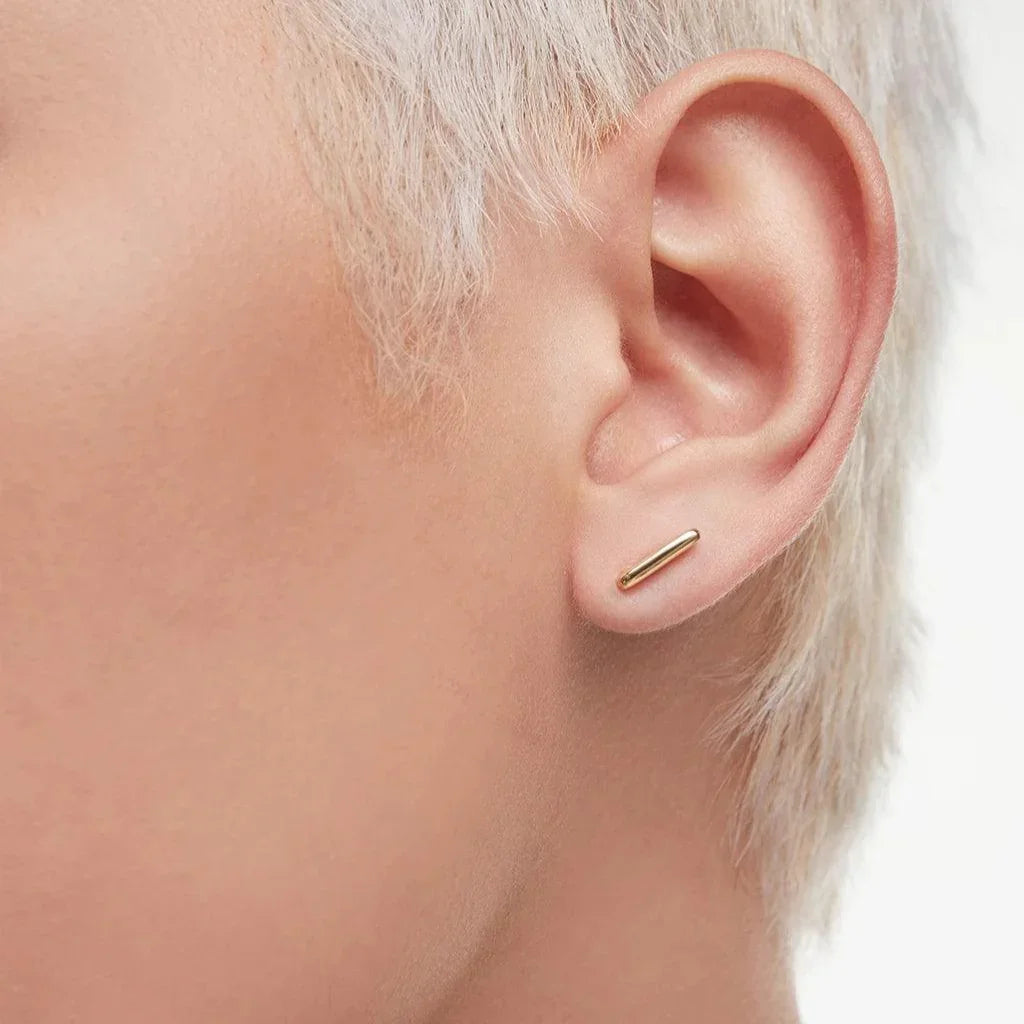 Close-up of a person with short platinum blonde hair wearing a minimalist, horizontal gold bar stud earring.