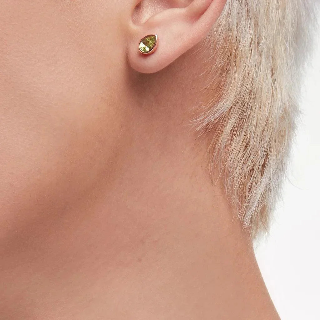 Close-up of a single ear wearing a minimalist pear-shaped green gemstone stud earring in a polished gold bezel setting.