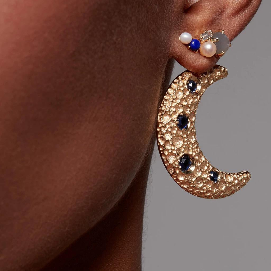 Gold crescent moon earring with blue gemstones worn on ear with multiple stones climber earring