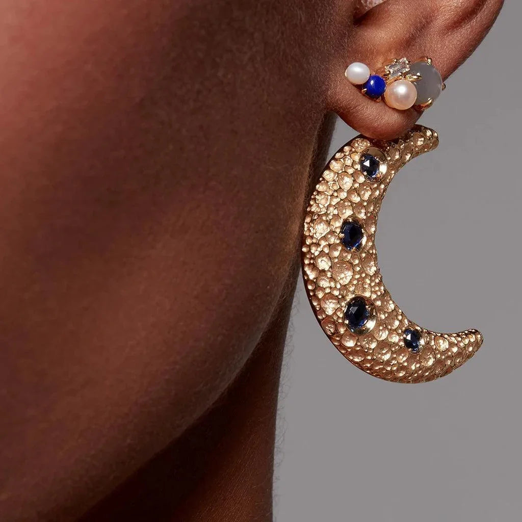 Gold crescent moon earring with blue gemstones worn on ear with multiple stones climber earring