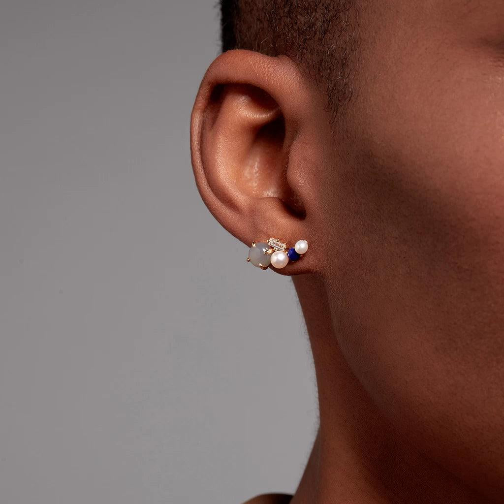 Close-up of silver cluster stud earring with pearls and blue gemstones on dark skin.