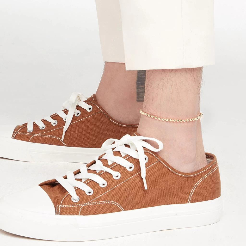 Men’s ankle adorned with a delicate gold bead anklet, paired with cream pants and brown canvas sneakers.