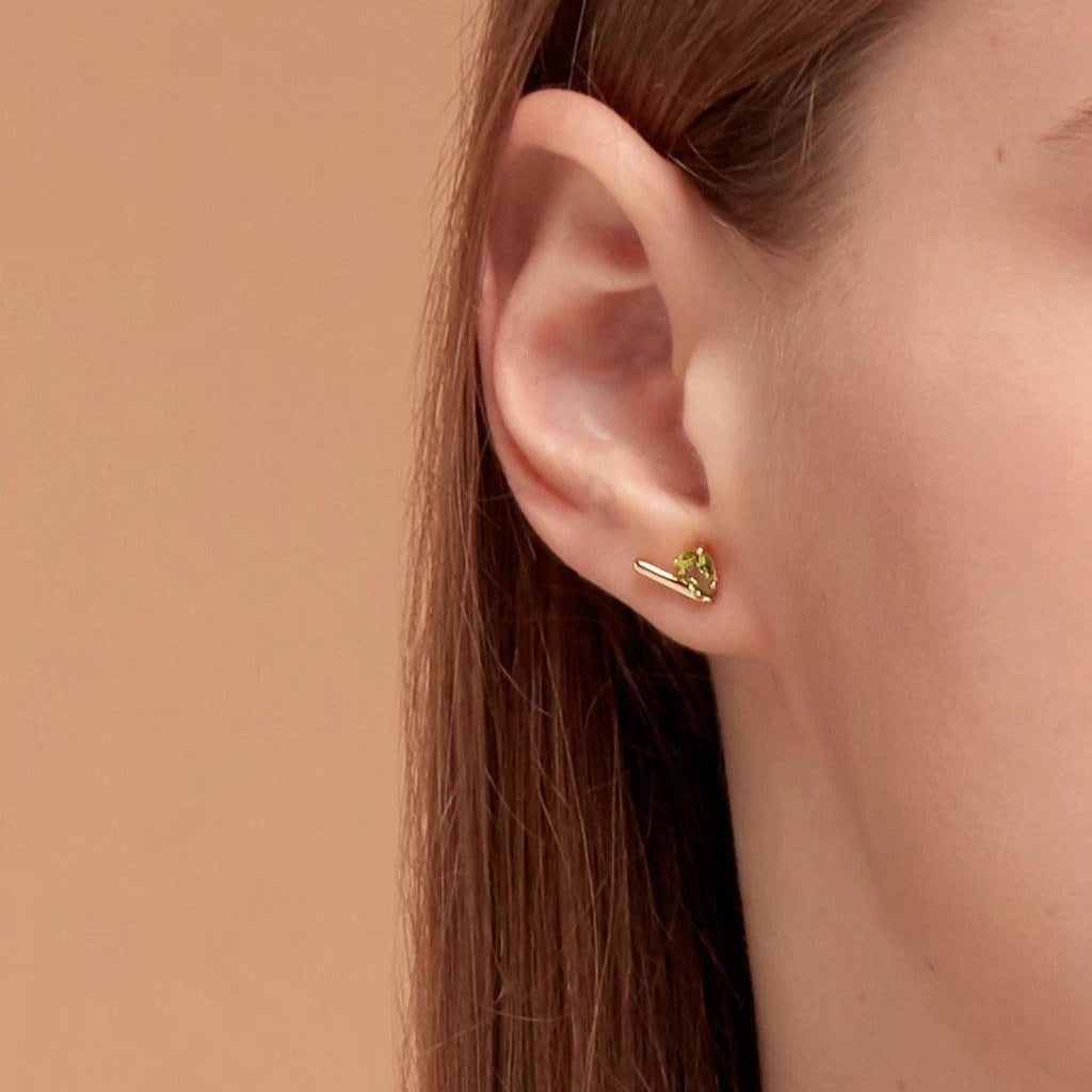 Close-up of a single ear wearing a minimalist gold bar stud with green peridot gemstone.