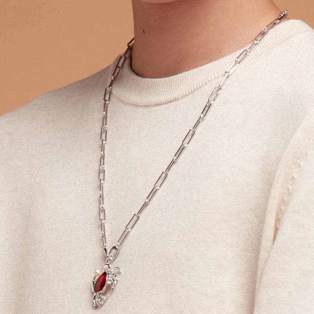 Long silver chain necklace with crab charm featuring a red stone and pearl, styled over a beige sweater.