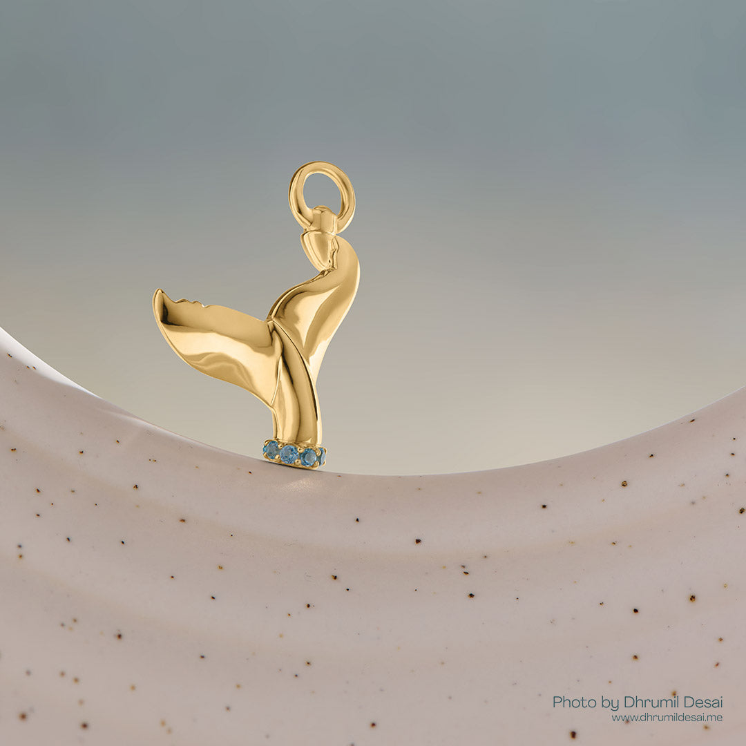 Gold whale tail charm with small blue gemstones at the base, displayed on a neutral curved surface.