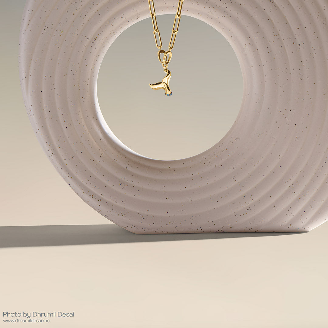 Gold paperclip chain necklace with a whale tail pendant hanging inside a speckled circular sculpture.