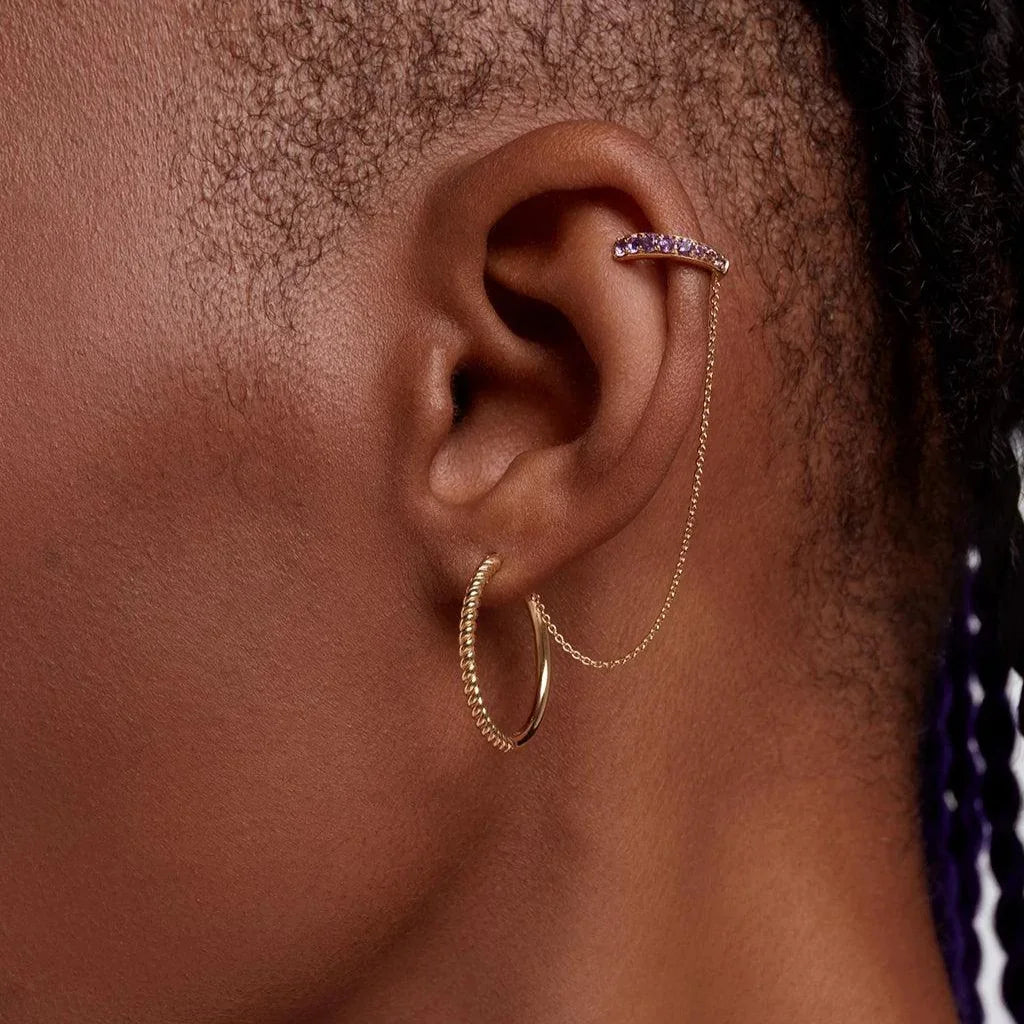 Close-up of gold ear cuff with purple gemstones and delicate chain on a dark-skinned ear, paired with a textured gold hoop.