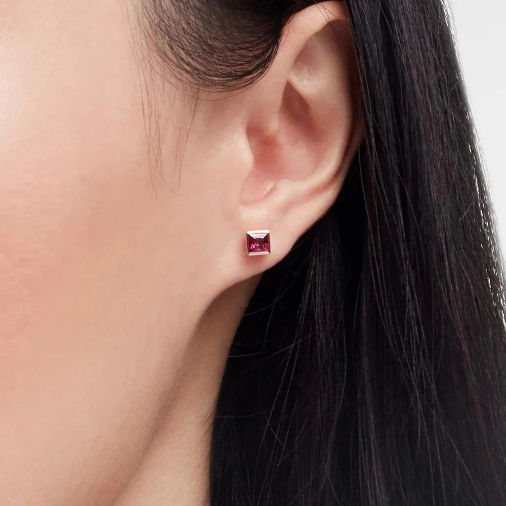Close-up of an ear wearing a single small square stud earring with a vivid ruby-red gemstone set in polished metal, showcased against smooth skin and straight dark hair.