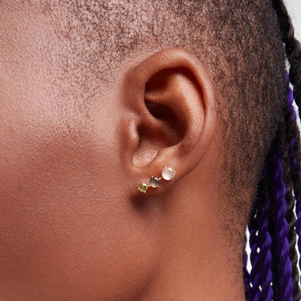 Close-up of an ear wearing a rose quartz, peridot, and grey pearl cluster stud earring with gold setting on dark skin.
