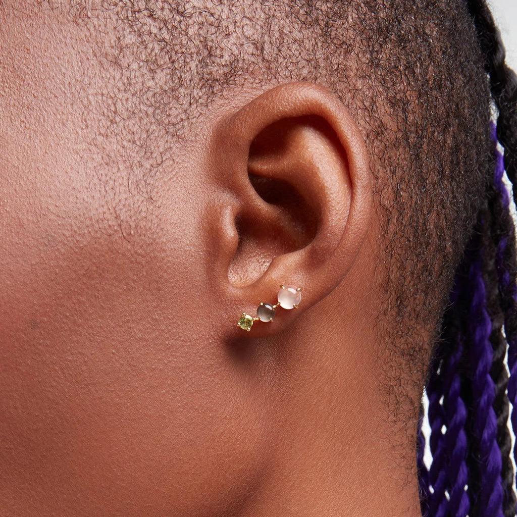 Close-up of an ear wearing a rose quartz, peridot, and grey pearl cluster stud earring with gold setting on dark skin.