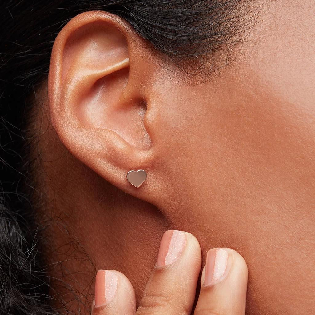 Close-up of an ear with a small rose gold heart-shaped stud earring, worn on the earlobe.