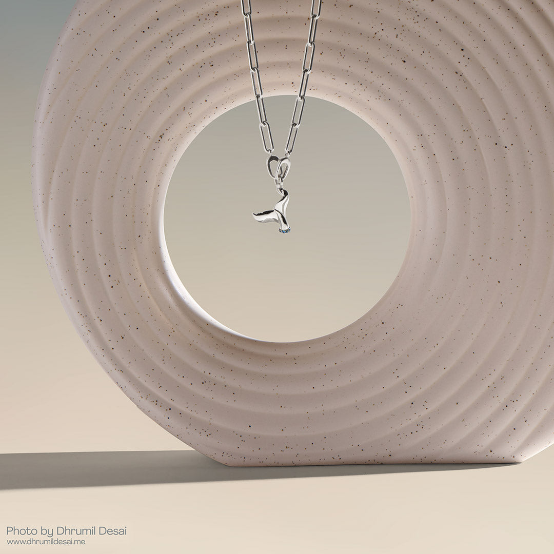 Silver paperclip chain necklace with a whale tail pendant displayed inside a speckled circular sculpture.