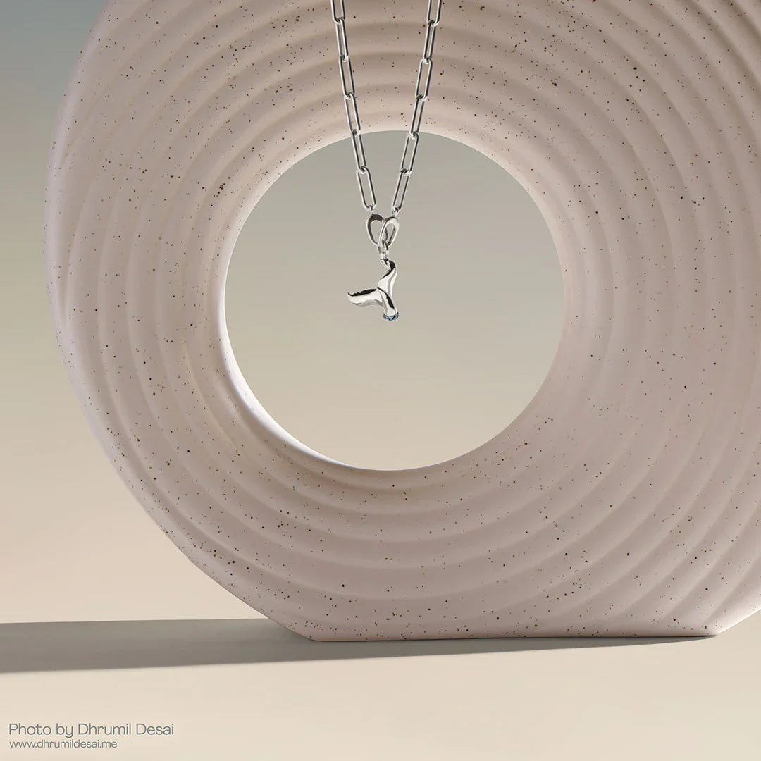 Silver paperclip chain necklace with a whale tail pendant displayed inside a speckled circular sculpture.