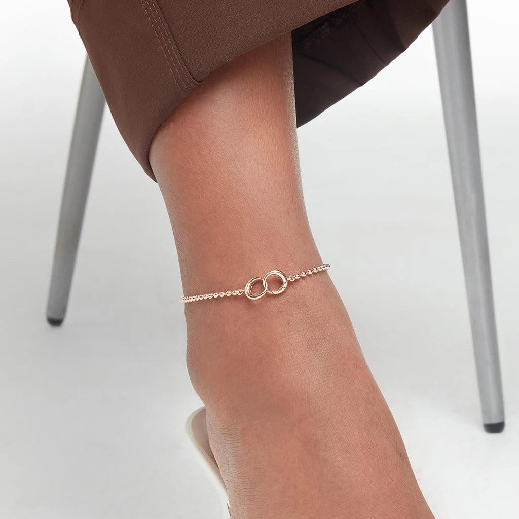 Minimal rose gold anklet with interlocking circles on a woman's ankle, styled with brown pants and white heels.