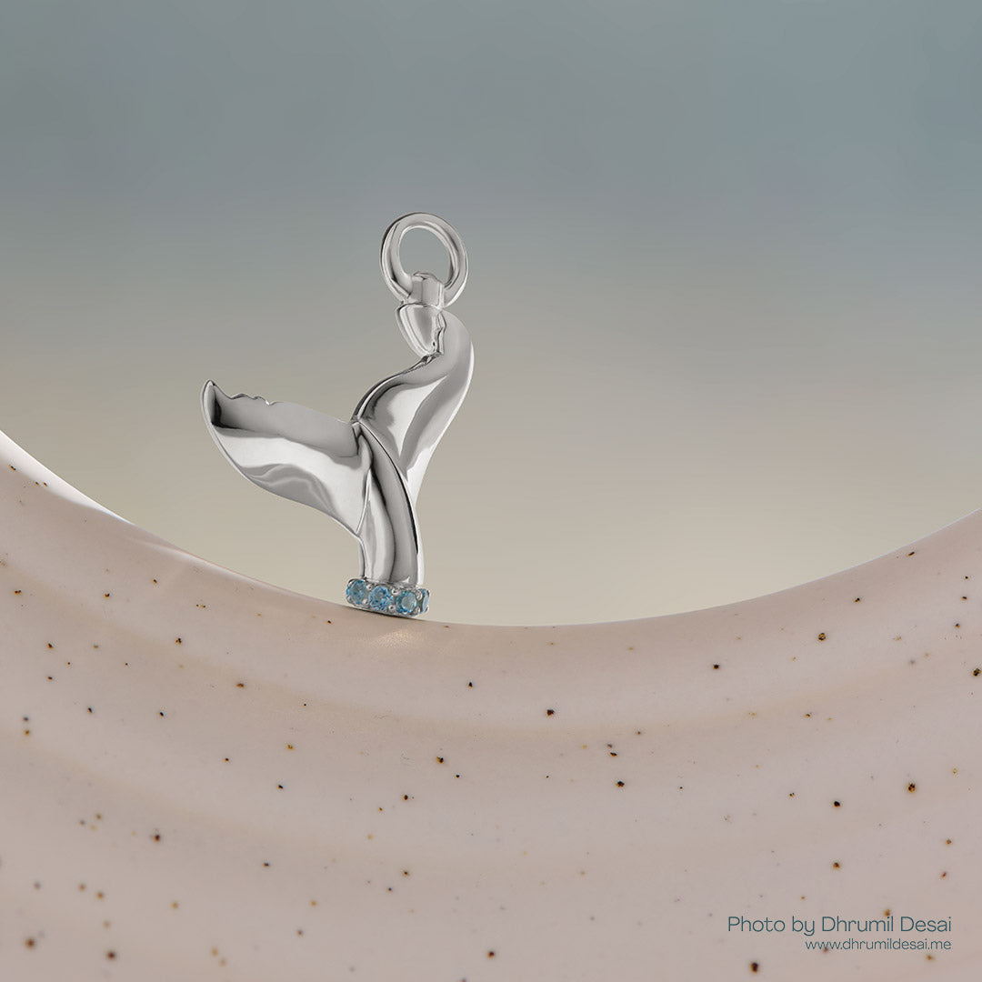 Silver whale tail charm with blue gemstones at the base displayed on a neutral curved surface.