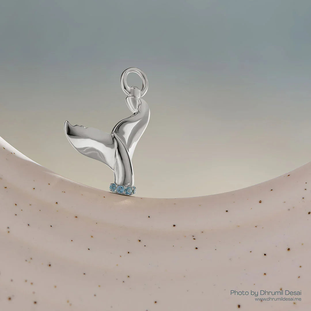 Silver whale tail charm with blue gemstones at the base displayed on a neutral curved surface.