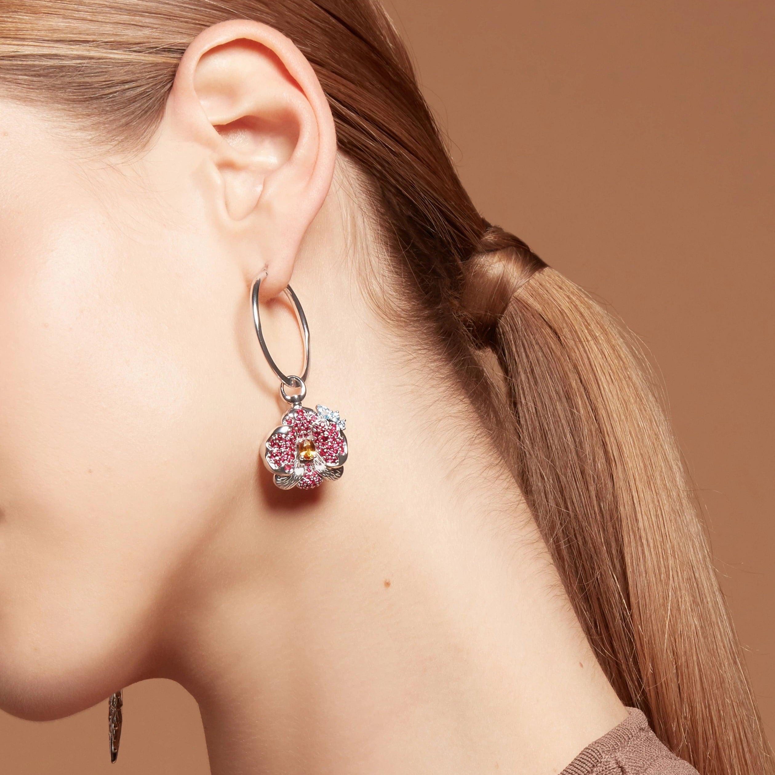 Close-up of a woman's ear wearing a silver round hoop earring adorned with a large pink orchid flower charm encrusted with gemstones.