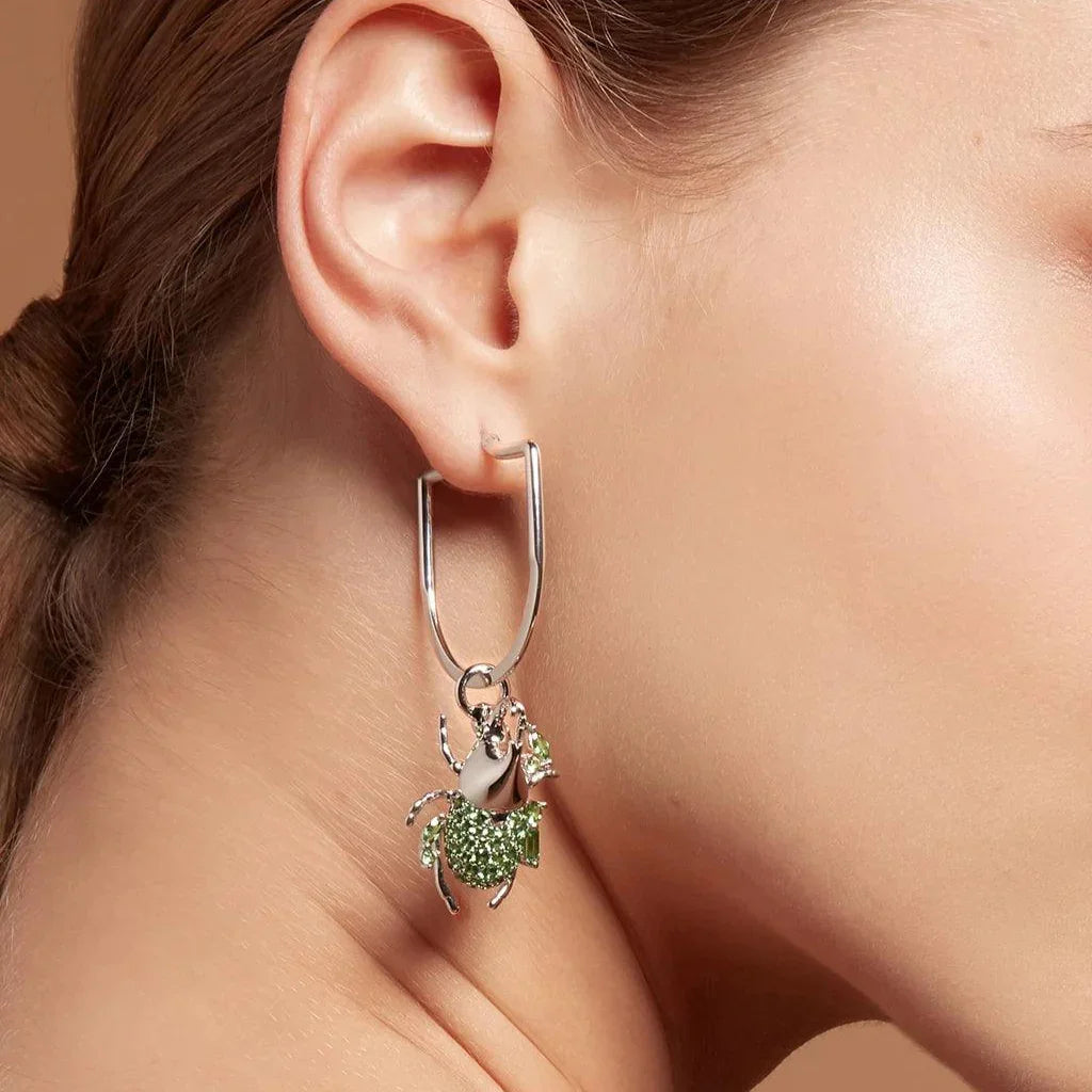 Close-up of a woman's ear wearing a polished silver hoop earring with a dangling green gemstone-encrusted beetle-shaped charm.