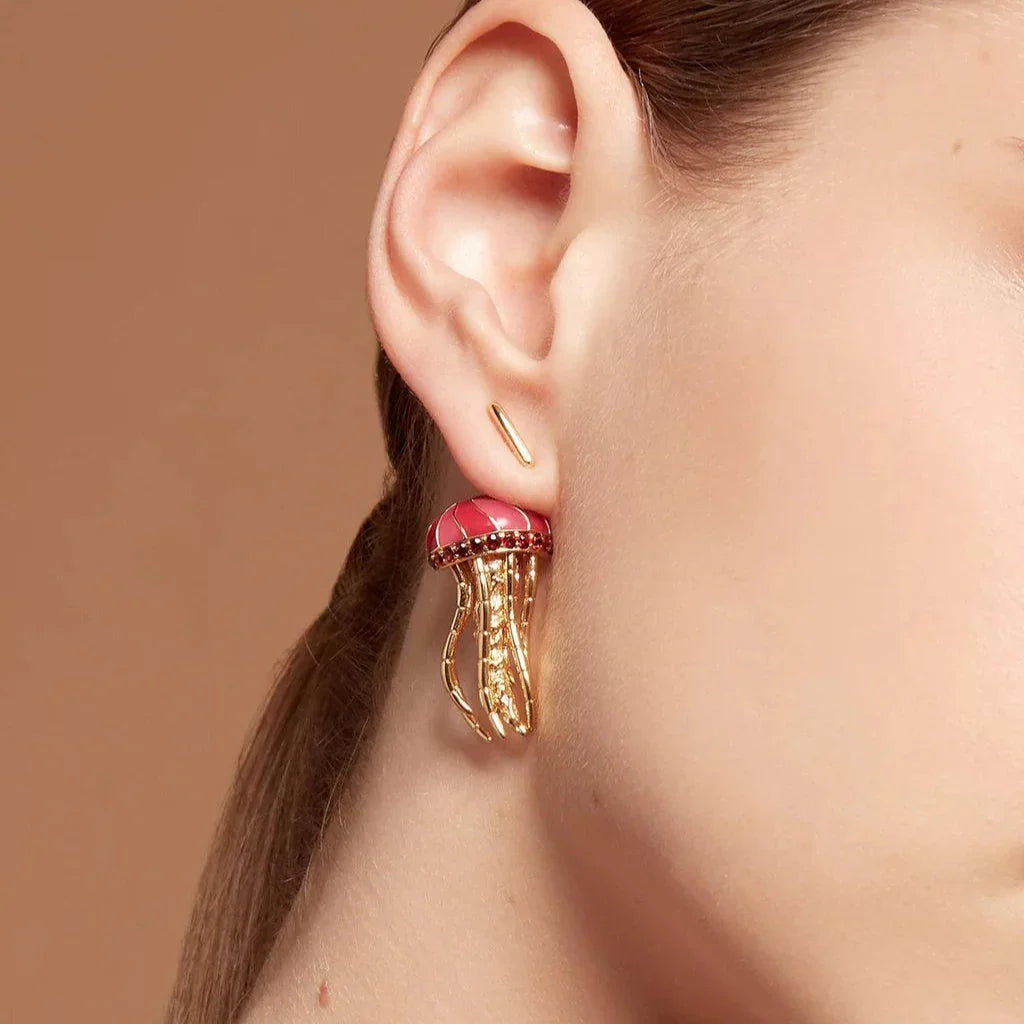 Close-up of a woman's ear wearing a gold and pink jellyfish-shaped earring with dangling tentacle-like details, styled with a sleek ponytail.