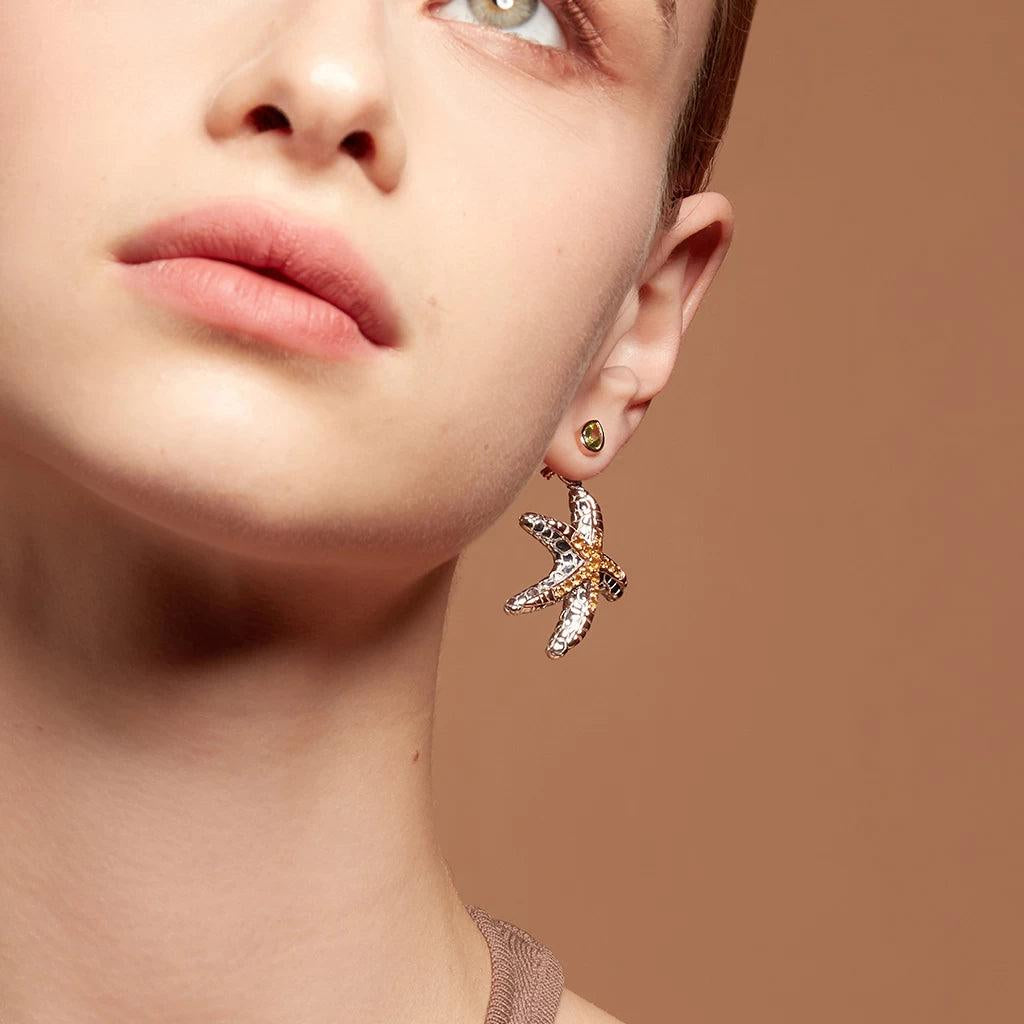 Woman wearing a pair of teardrop-shaped green gemstone stud earrings with a gold bezel setting, styled with a large starfish-shaped drop charm attached below.
