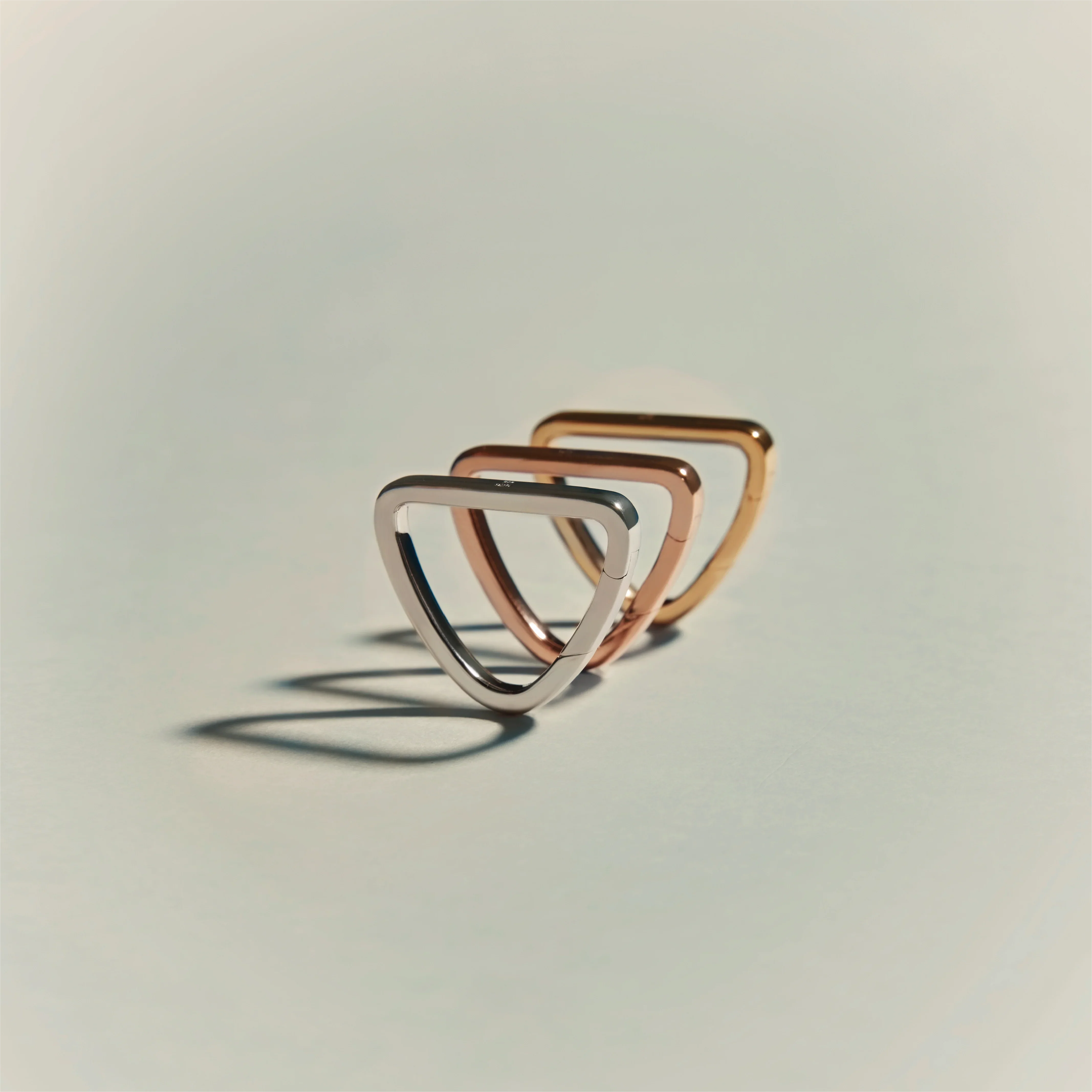 Three geometric triangular rings in silver, rose gold, and yellow gold arranged in a row on a light surface.