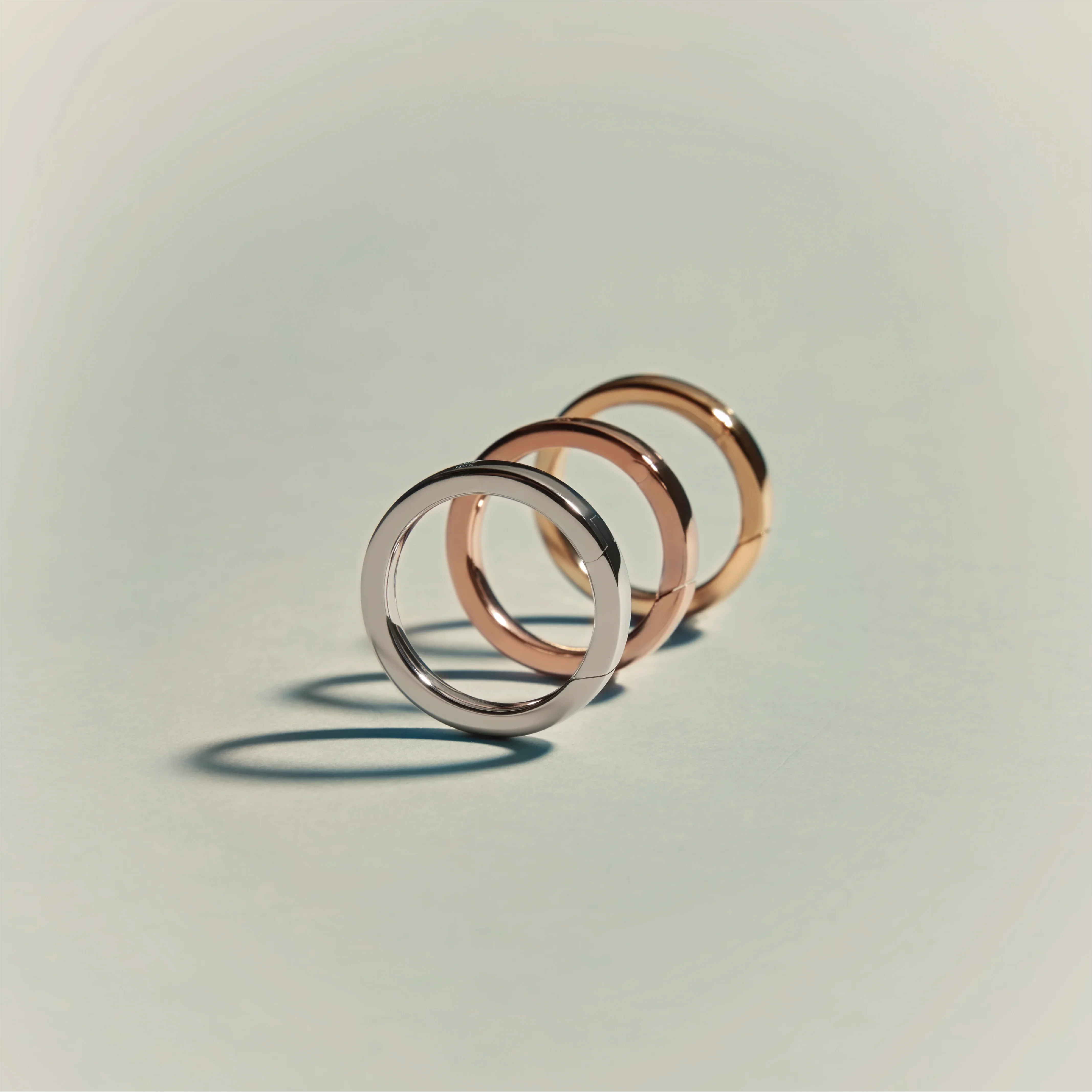 Three minimalist circular rings in silver, rose gold, and yellow gold finishes arranged in a row on a light surface.