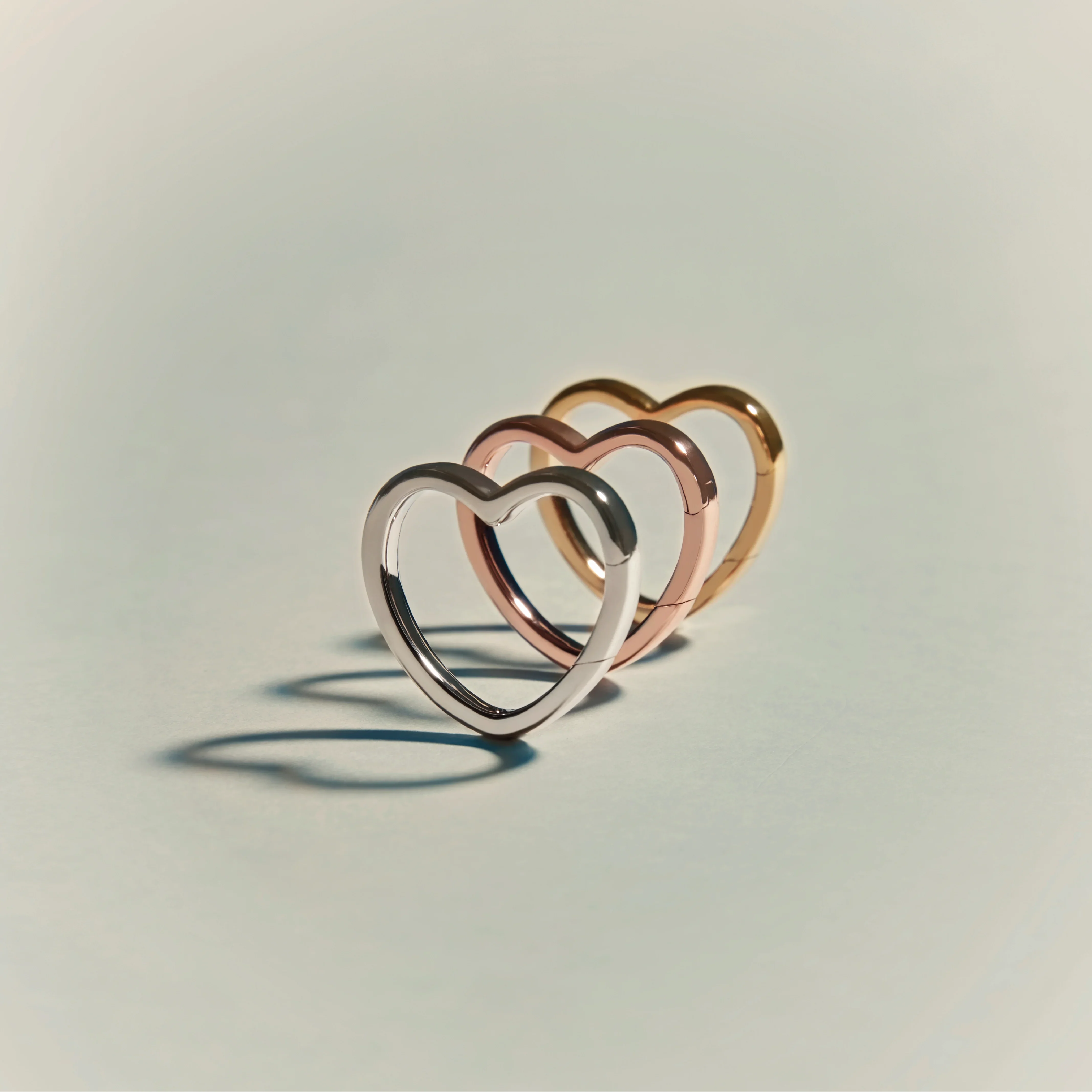 Three heart-shaped rings in silver, rose gold, and yellow gold finishes arranged in a row on a neutral surface.