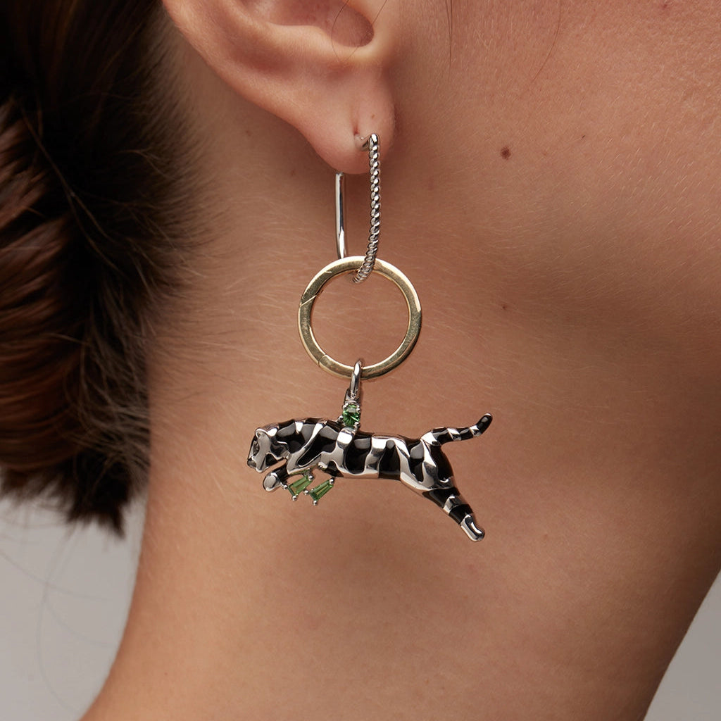 Close-up of a silver and gold hoop earring featuring a black and white enamel panther charm with small green gemstone accents, worn on ear.