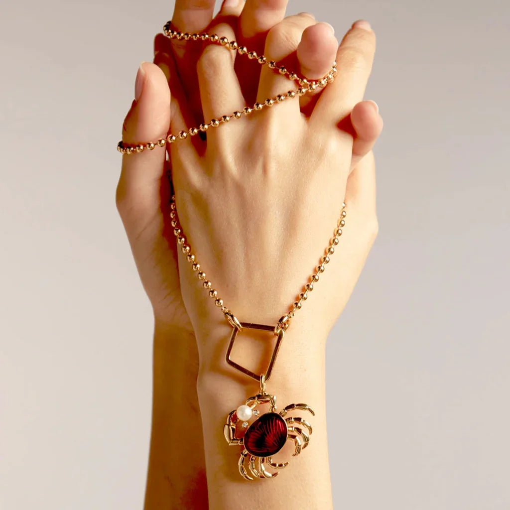 Hands wrapped in gold ball chain necklace featuring a red enamel crab charm with a pearl accent.