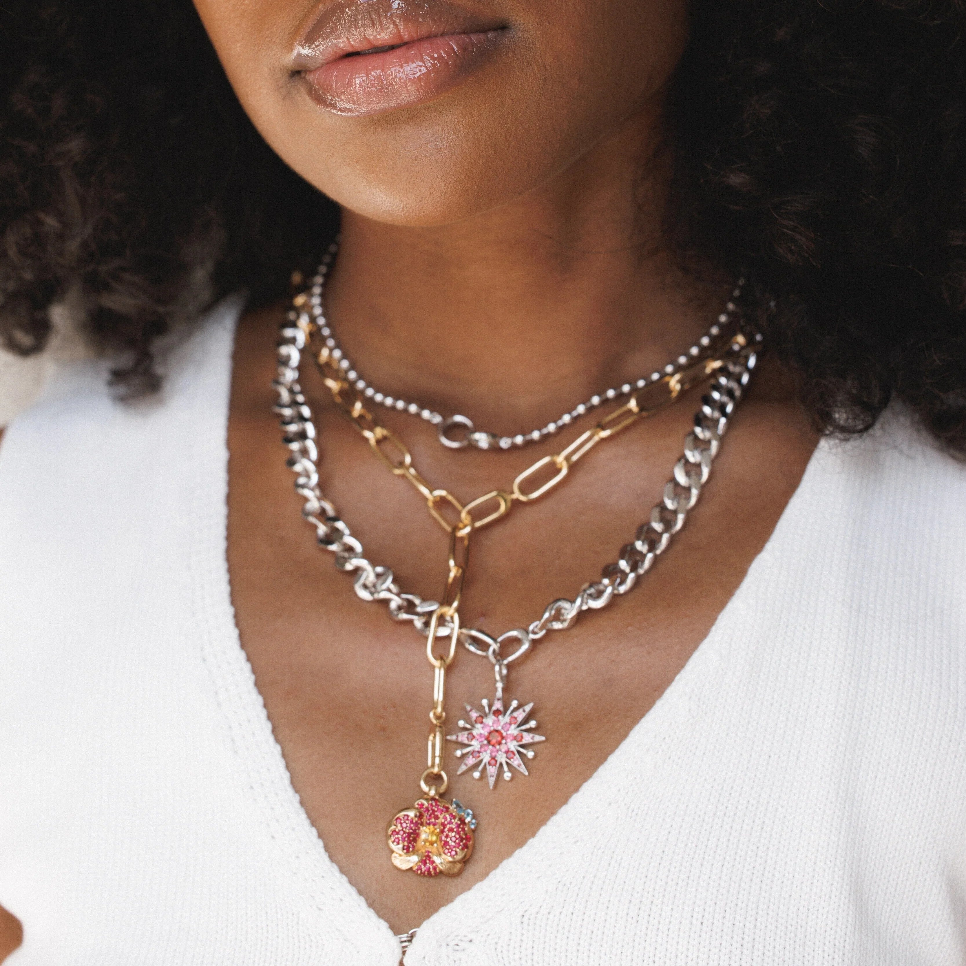 Close-up of a woman wearing layered mixed-metal necklaces with large chain links in silver and gold. Two charms hang at the longest layer: a gold orchid-shaped charm set with pink gemstones, and a silver starburst pendant with pink and red stones.
